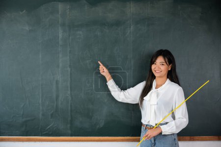 Foto de Back to school concept. Happy beautiful young woman standing hold pointer to back board, Asian female teacher smiling with wooden stick pointing to blackboard at school in classroom, Education - Imagen libre de derechos