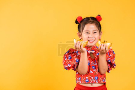 Foto de Año Nuevo Chino. Feliz asiática china niña sonrisa usando cheongsam rojo sosteniendo lingote de oro, niños retrato en vestido tradicional celebrar barra de oro, estudio corto aislado sobre fondo amarillo - Imagen libre de derechos