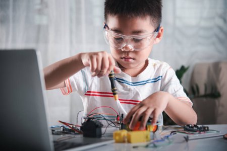 Foto de Niño asiático que ensambla el proyecto de la tarea del coche del robot de Arduino en el hogar, niño pequeño aprieta la tuerca con un destornillador para montar el juguete del coche, creando la lección electrónica de la escuela del taller de la tecnología de AI - Imagen libre de derechos