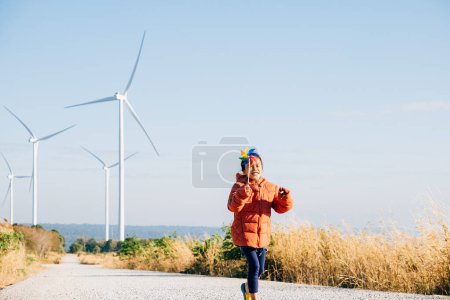 Foto de Pequeñas niñas felicidad cerca de molinos de viento que sostienen molinos encarna la educación de energía eólica juguetona. Belleza de electricitys limpia exhibida en un ambiente alegre de turbina de viento soleado. - Imagen libre de derechos