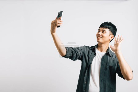 Photo for Portrait of a cheerful Asian young man taking a selfie with his smartphone, capturing his excitement in a studio shot isolated on white. Fun with photography - Royalty Free Image