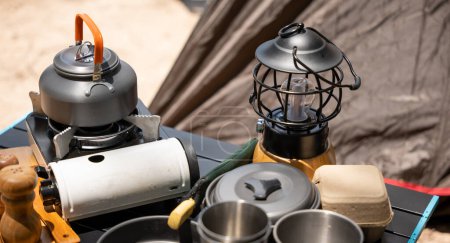 Photo for Cooking gear for a tranquil beach campsite, kettle, pot, pan, gas stove, flashlight, and camera set on a table by the tent. Enjoy a serene camping experience amidst nature. - Royalty Free Image