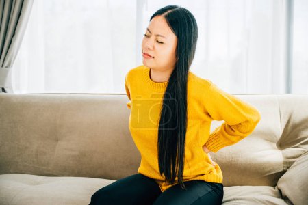 Photo for An Asian woman sitting on sofa holds her lower back in unbearable pain. Depicting chronic back pain issues discomfort and the need for medical care and attention. healthcare and problem concept - Royalty Free Image