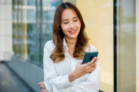 Photo for An Asian girl is enjoying the outdoors while using her smartphone. She is swiping and typing on the device. The woman is connected and engaged. - Royalty Free Image