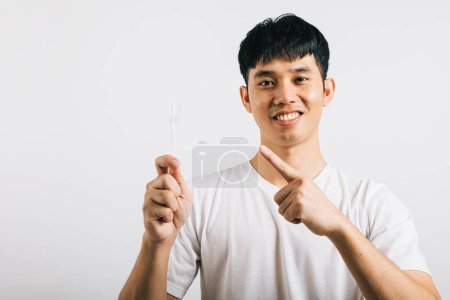 Foto de Sonriente joven asiático, en un concepto de salud dental, cepilla los dientes con un cepillo de dientes y señala con entusiasmo. Estudio aislado sobre fondo blanco, mostrando una actitud positiva. - Imagen libre de derechos