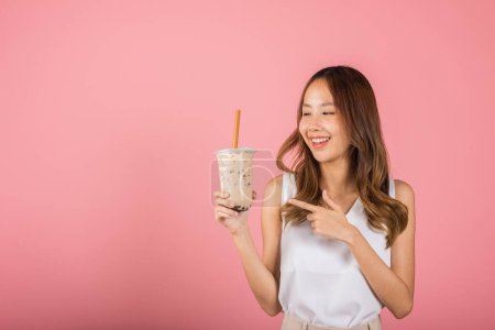 Téléchargez les photos : Asian beautiful young woman holding drinking brown sugar flavored tapioca pearl bubble milk tea and pointing finger, smile portrait female, isolated on pink background, pearl milk tea beverage concept - en image libre de droit