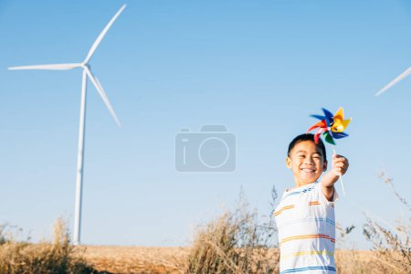 Foto de El niño alegre que juega con el juguete del molinete cerca de los aerogeneradores celebra el espíritu de la energía eólica. Muestra la innovación en electricidad limpia en medio de una tranquila granja de molinos de viento en el campo contra un cielo azul brillante. - Imagen libre de derechos