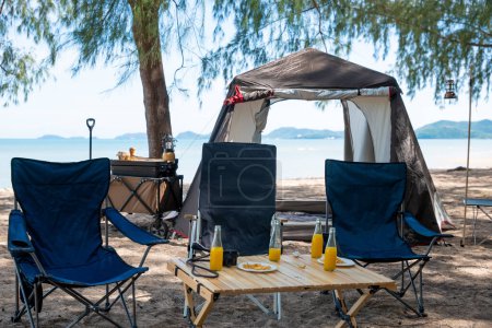 Foto de Relájese junto al mar, sillas, mesa de picnic y tienda de campaña crean la experiencia de camping perfecta. Puesta de sol, familia, y el gran aire libre aquí es donde comienza el viaje a la naturaleza. El medio ambiente está llamando. - Imagen libre de derechos