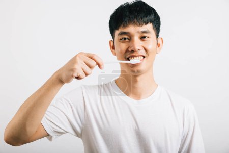 Téléchargez les photos : Portrait d'un jeune Thaïlandais souriant se brossant les dents avec confiance pour la santé buccodentaire. Studio tourné isolé sur fond blanc, favorisant les soins dentaires et buccodentaires. - en image libre de droit