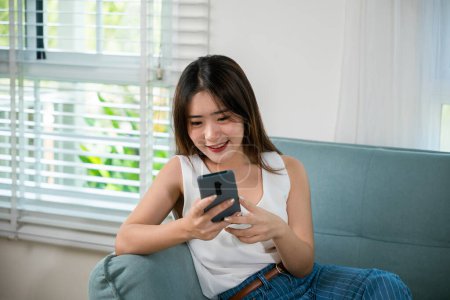 Téléchargez les photos : Social media networks. Happy beautiful female relax and shopping online with smartphone, Smiling Asian woman sitting on sofa reading news on mobile smart phone at living room at home - en image libre de droit