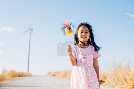 Foto de Una niña alegre sonriendo y corriendo con molinos cerca de molinos de viento. Abrazar la energía limpia a través de una educación lúdica en un pintoresco entorno de turbina eólica bajo un hermoso cielo. - Imagen libre de derechos