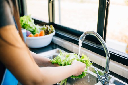 Photo for In a modern kitchen a woman washes fresh vegetables under running water in the sink preparing a vegan salad. Emphasizing cleanliness and freshness in homemade healthy food. - Royalty Free Image
