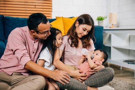 Photo for A loving mother sits in the living room feeding her newborn baby with a milk bottle while her toddler son and daughter bond with dad. Family togetherness is the heart of joy at home. - Royalty Free Image