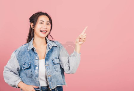Téléchargez les photos : Heureux portrait asiatique belle jeune femme mignonne debout souriant indiquer doigt vide espace regardant à la caméra, studio tourné isolé sur fond rose, Thai femelle pointant index avec espace de copie - en image libre de droit