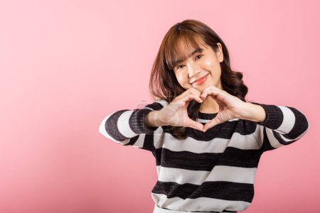 Photo for Portrait of a happy Asian woman with a confident smile, making a heart symbol with her fingers and hands on a pink background. Sending love and joy for Valentines Day. - Royalty Free Image