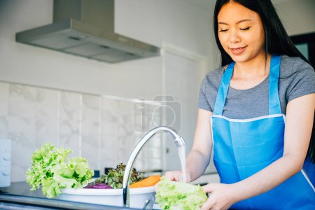 Photo for A smiling woman washes vegetables in the kitchen sink readying them for a delicious salad. Emphasizing hygiene and her passion for healthy eating in her home. - Royalty Free Image