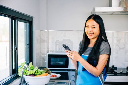 Photo for In a cozy kitchen an Asian woman cooks vegetables viewing a cooking tutorial on her smartphone. Smiling and inspecting fruits she merges modern tech with culinary exploration. - Royalty Free Image