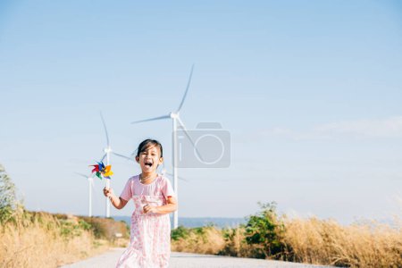 Foto de Niñas felicidad visto cerca de molinos de viento sosteniendo molinos mientras se ejecuta. Visuales lúdicos de educación en energía eólica que muestran la generación de electricidad limpia bajo un cielo sereno. - Imagen libre de derechos