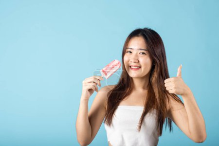 Photo for Sweet tasty frozen dessert icecream on summer time. Happy Asian young beautiful woman holding delicious ice cream wood stick mixed fruit flavor and show thumb up good sign, isolated on blue background - Royalty Free Image