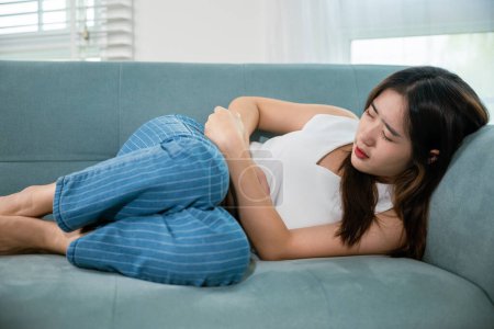 Photo for Sad Asian woman close eyes she problem painful stomachache, young female unhappy hands holding on stomach suffering from abdominal pain on sofa at home in living room, medical care and health concept - Royalty Free Image