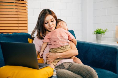 Téléchargez les photos : Équilibrer la maternité et le travail d'une femme d'affaires types sur son ordinateur portable que sa fille bébé fait une sieste paisible sur le canapé. Ce portrait capture leur lien d'amour. - en image libre de droit