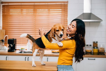 Foto de En la acogedora cocina, una mujer asiática sonriente encuentra alegría jugando con su perro Beagle. Su vínculo muestra la felicidad, la unión y la amistad propietario-mascota que traen diversión a la familia. Amor de mascotas - Imagen libre de derechos