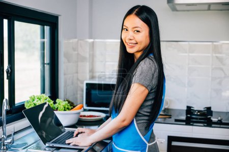 Photo for In the kitchen a smiling woman stands engrossed in cooking and using her laptop. Emphasizing the joy of technology in aiding culinary experiments and learning new recipes. - Royalty Free Image