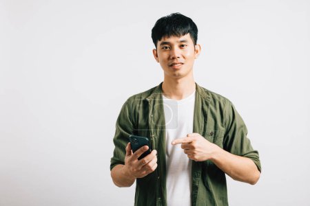 Photo for Smiling Asian man confidently points at his smartphone screen for online shopping. Studio portrait isolated on white, highlighting his elegant use of technology. Copy space is evident. - Royalty Free Image