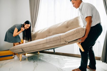 Téléchargez les photos : Portrait d'un couple asiatique souriant déplaçant joyeusement un canapé dans leur nouvel appartement. Idéal pour représenter l'excitation de placement de meubles et de tassement de la maison. Jour de déménagement - en image libre de droit
