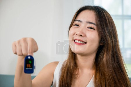 Foto de Mujer joven asiática que utiliza equipo de oxímetro médico para medir la saturación de oxígeno de pulso en el sofá en la sala de estar en el hogar, Auto salud con pandemia de virus corona, atención médica - Imagen libre de derechos