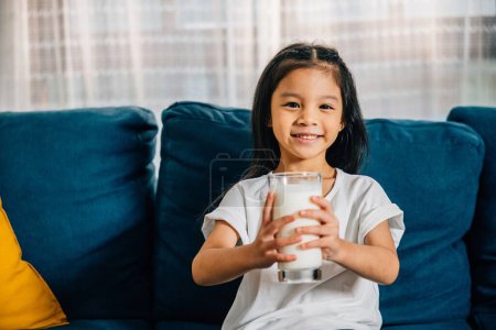 Téléchargez les photos : Gros plan d'une petite fille asiatique tenant un verre de lait assis sur le canapé à la maison. Cette image charmante représente l'importance de la nutrition des soins de santé et la joie de l'enfance. - en image libre de droit