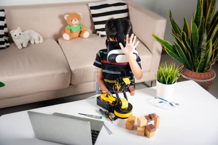 Téléchargez les photos : Simulateur de réalité virtuelle. Enfant asiatique utilisant des lunettes VR sur bras robotisé en atelier, enfant apprenant programmeur contrôle bras robot avec capteurs pour ramasser bloc de bois, éducation technologique. industrie 4.0 - en image libre de droit
