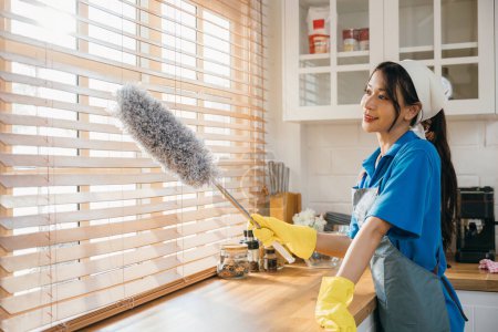 Photo for Smiling Asian woman holding duster cleans dirty window blinds. Ensuring purity and hygiene in routine housework. Modern cleaning service for a clean hom. whisk - Royalty Free Image