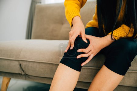 Téléchargez les photos : Soins de santé et concept médical, Une jeune femme tient son genou douloureux sur un canapé représentant l'arthrite tendineuse chronique. Mettre en évidence la douleur et l'inflammation des tendons. - en image libre de droit
