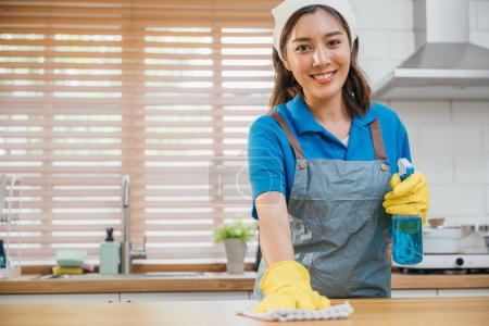 Photo for Asian woman in yellow gloves cleans wooden kitchen counter with liquid spray ensuring hygiene. Housekeeping service focusing on home cleanliness. Clean disinfect home care. maid household job. - Royalty Free Image
