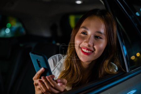 Foto de Feliz hermosa mujer mensajes de texto teléfono inteligente sentado asiento trasero del coche en la ciudad, mujer de negocios asiática que trabaja hasta tarde viajando desde la oficina en el asiento trasero del taxi con el teléfono móvil en la ciudad por la noche después del trabajo tardío - Imagen libre de derechos