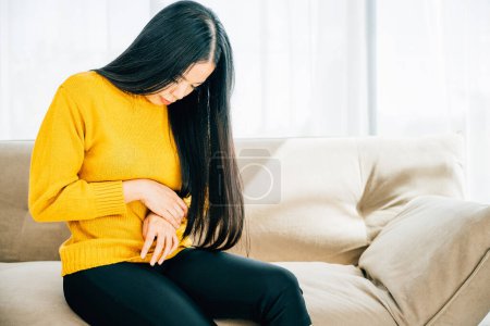 Photo for Highlighting stomachache, Asian woman on home couch endures abdominal pain. Depicting discomfort symptoms and the importance of medical care for stomachache. Health care abdomen bloating concept - Royalty Free Image