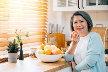 Photo for A smiling old Asian woman, a grandmother, finds joy in the kitchen, cooking and preparing healthy vegan food. She selects a fresh apple for eating, emphasizing the diet concept and happiness. - Royalty Free Image