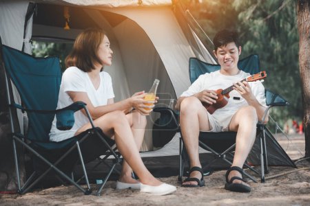 Foto de Momentos llenos de amor en el campamento, Una pareja asiática tocando el ukelele y cantando, rodeada por el calor de su tienda de campaña. Sus sonrisas revelan la alegría de estar juntos. La felicidad está en el aire. - Imagen libre de derechos