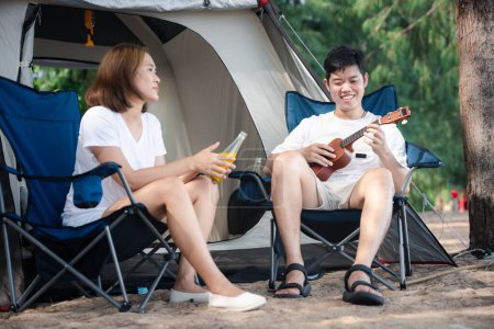 Téléchargez les photos : Camping bliss, Un couple asiatique joyeux, par leur tente, sérénade l'un l'autre avec des airs ukulélés, répandant l'amour et sourit sous le soleil couchant. Ensemble, c'est leur mélodie. Camping extérieur - en image libre de droit