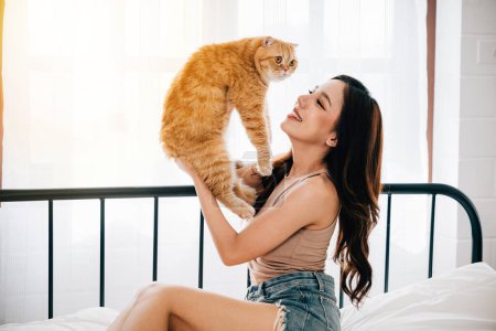 Photo for A close-up portrait of a cheerful young woman at home, holding her cute orange Scottish Fold cat with affectionate satisfaction in the bedroom. A heartwarming display of friendship and joy. Pet love - Royalty Free Image