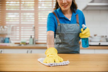 Téléchargez les photos : Asiatique nettoyage professionnel portant des gants jaunes essuie comptoir de cuisine en bois avec pulvérisation liquide. Le ménage à la maison met l'accent sur la propreté. Nettoyer désinfecter les soins à domicile. femme de ménage emploi. - en image libre de droit