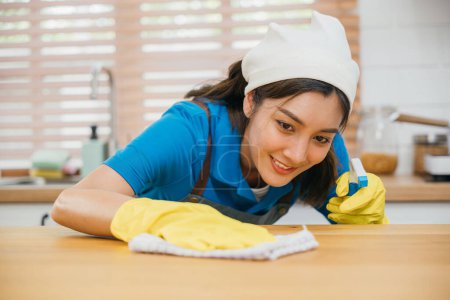 Téléchargez les photos : Femme asiatique en gants jaunes effectue le nettoyage ménager comptoir de cuisine avec pulvérisation liquide. Mettre l'accent sur la propreté et l'hygiène de la maison. Nettoyer désinfecter les soins à domicile. femme de ménage emploi. - en image libre de droit