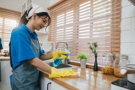 Photo for Employee in uniform cleaning kitchen desk with spray ensuring hygiene. Housekeeping and safety concept for a clean home. Maids diligent routine for home purity. Clean disinfect home care. Give me. - Royalty Free Image