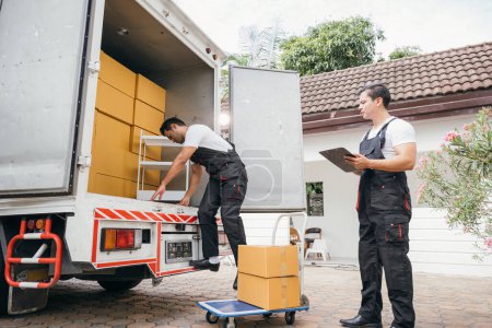Photo for Two employees of a removal company work together unloading furniture and boxes from the truck ensuring an efficient delivery into the new family home. Their teamwork delivers happiness. Moving Day - Royalty Free Image
