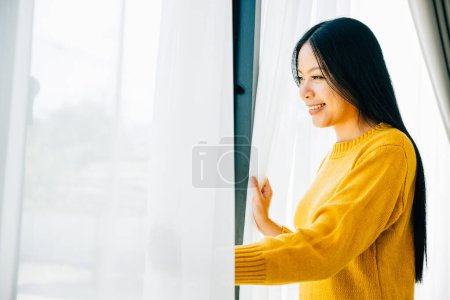 Photo for A young woman opens curtains in the early morning smiles at the view feeling relaxed and joyful at home. Embracing morning happiness relaxation and a cheerful start. - Royalty Free Image