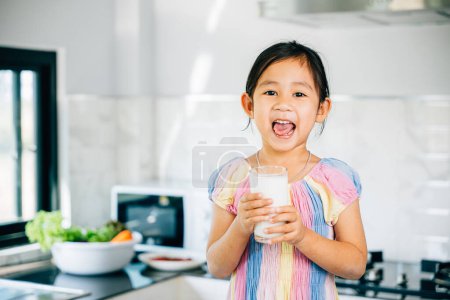 Photo for Adorable Asian preschooler holds milk cup in kitchen. Portrait of cute daughter enjoying drink smiling happily. Happy little girl relishing calcium-rich liquid radiating joy at home give me. - Royalty Free Image