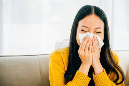Foto de Una mujer asiática sonándose la nariz sosteniendo un pañuelo lidiando con síntomas de gripe en su casa. Retrato de una mujer sintiéndose mal con mocos y tejido en la mano. - Imagen libre de derechos