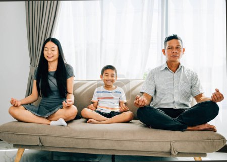 Téléchargez les photos : Les liens familiaux sereins, les parents et leur petit fils pratiquent le yoga assis dans une pose de lotus sur un canapé. Encourager l'harmonie équilibre et la tranquillité au sein de la maison. - en image libre de droit