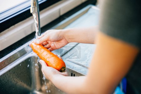 Photo for A young woman meticulously washes carrots in the kitchen sink emphasizing hygiene and organic food preparation. Illustrating the concept of washing vegetables before cooking. - Royalty Free Image